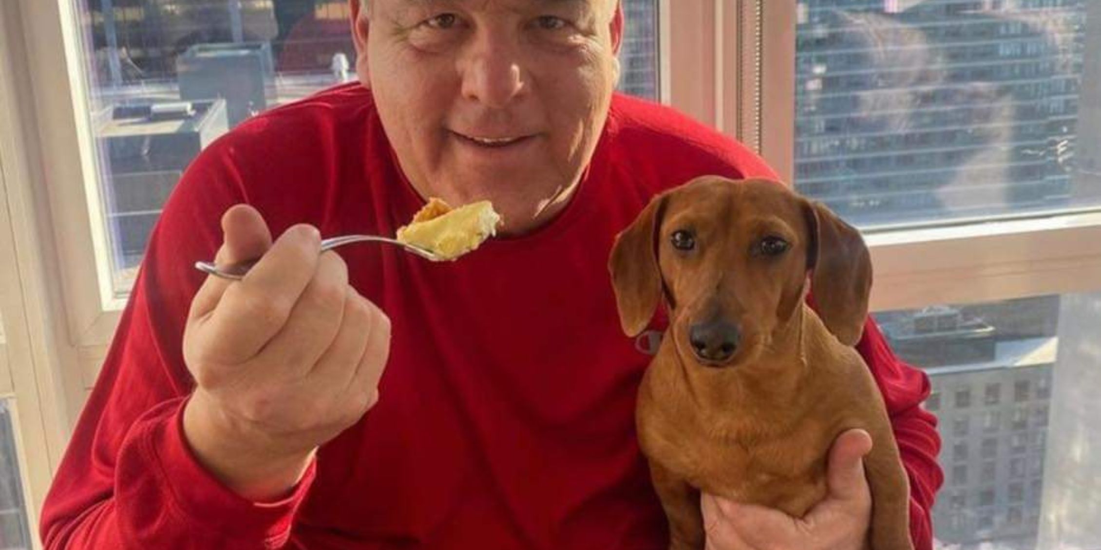 Blue Bloods BTS photo of Steve Schirripa in a red sweater holding Willie Boy while he eats cereal