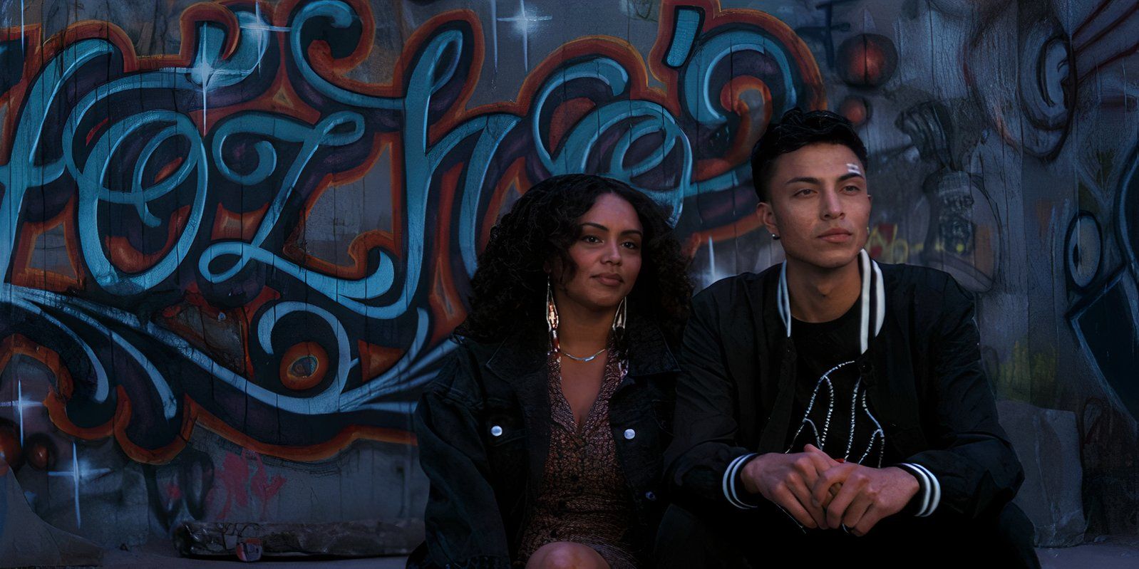 Krista (Zoey Reyes) and Jimmy Holiday (Kauchani Bratt) sitting in front of a wall of graffiti in Rez Ball