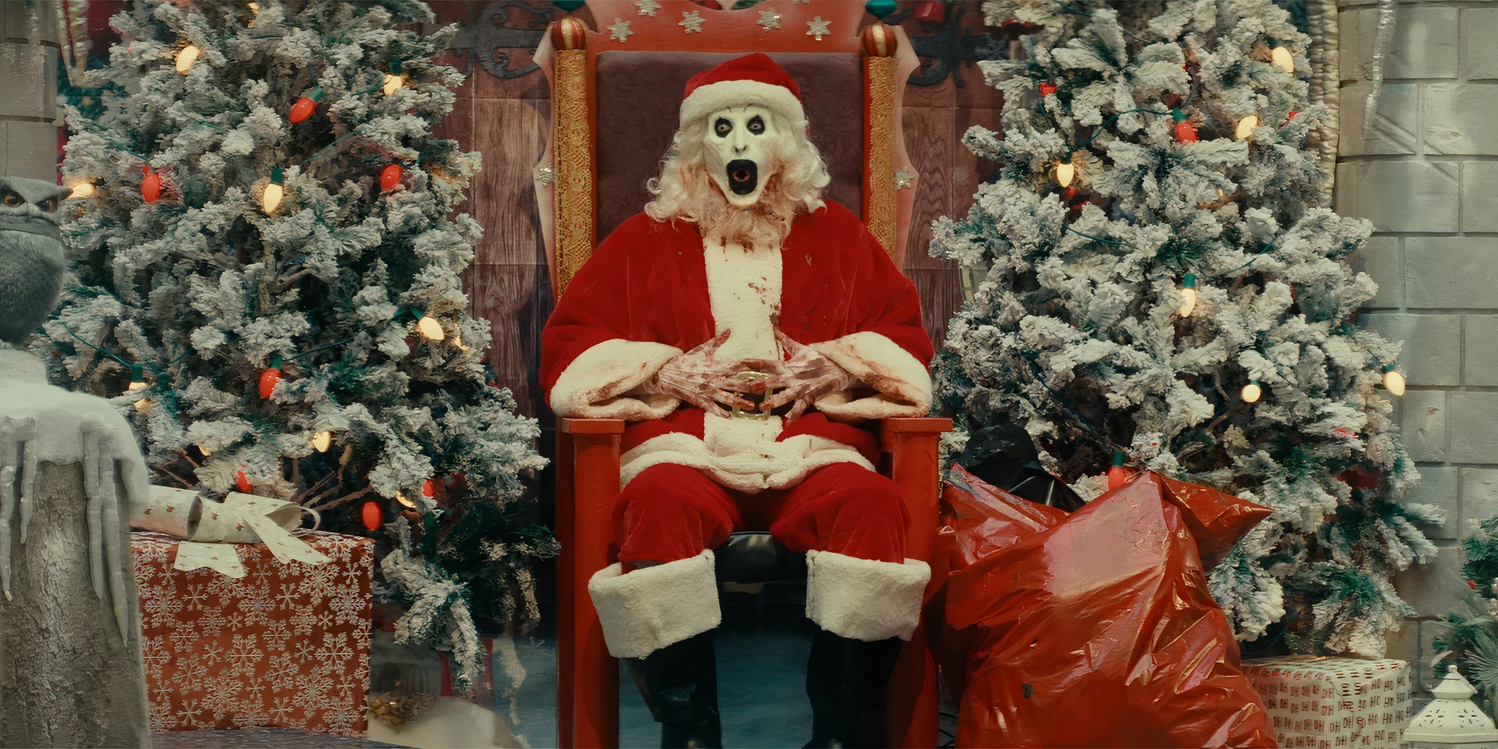 David Howard Thornton as Art the Clown making a silly face in a Santa Claus suit while sitting in a Santa mall exhibit in Terrifier 3