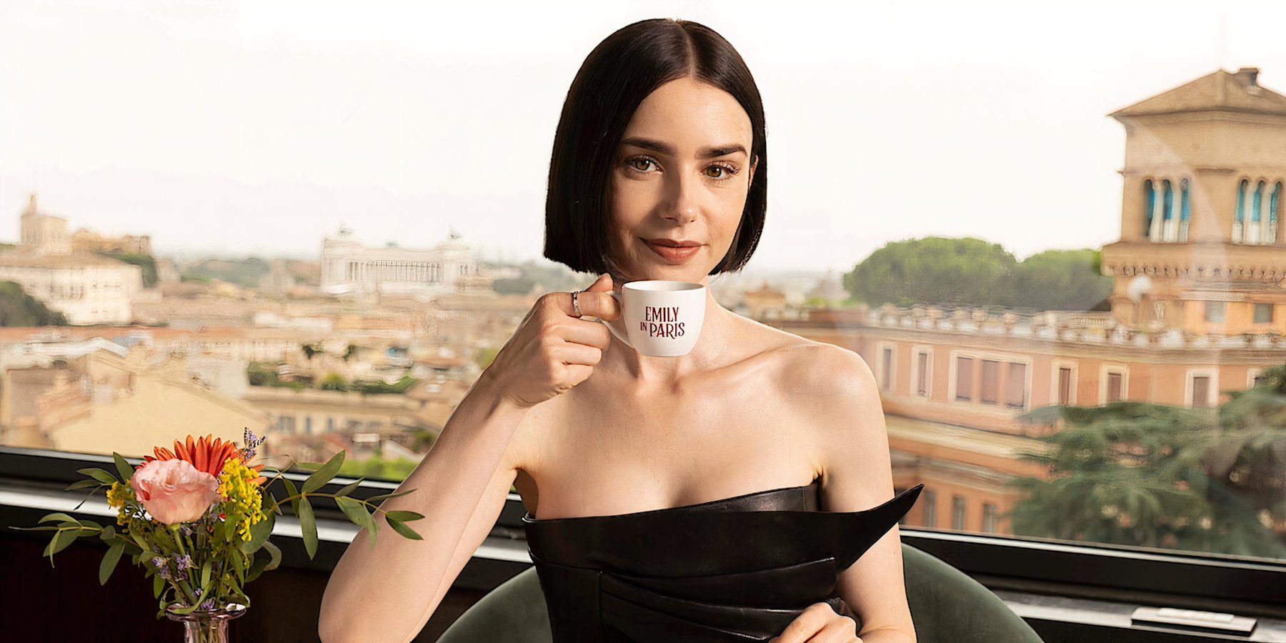 Emily in Paris's main character Emily sits at an outdoor cafe with a Paris backdrop. She holds a white teacup with the words' 