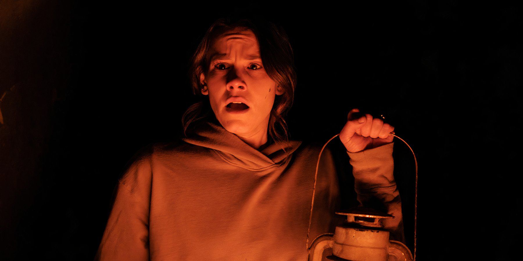 Smile 2022 protagonist Rose (Sosie Bacon) looks terrified holding up a lantern to partially light a pitch black room. 