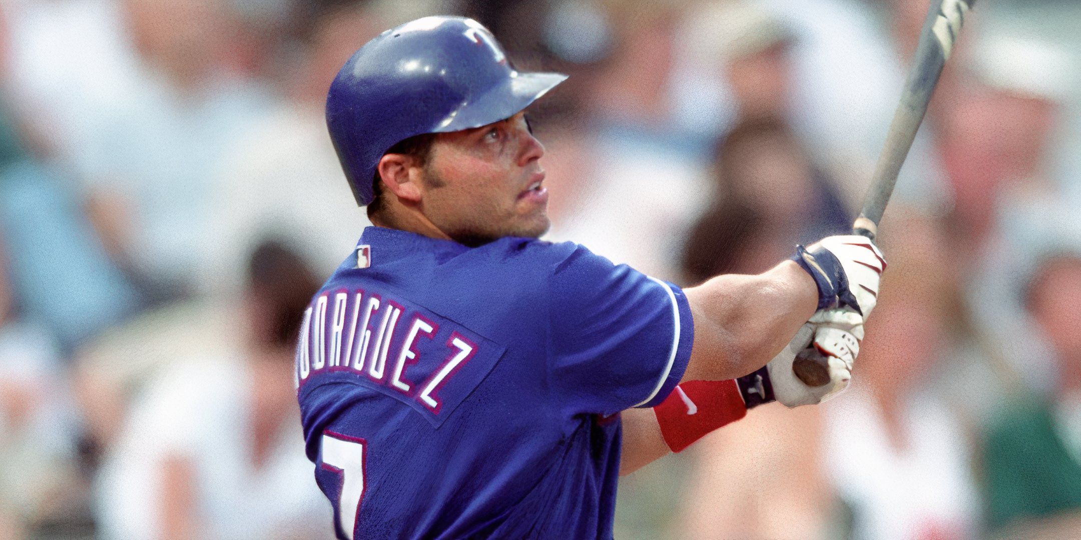 Iván Rodríguez, vestindo uniforme e capacete azul do Texas Rangers, é capturado no meio de um jogo. Seu nome, 'Rodríguez', está visível nas costas de sua camisa.