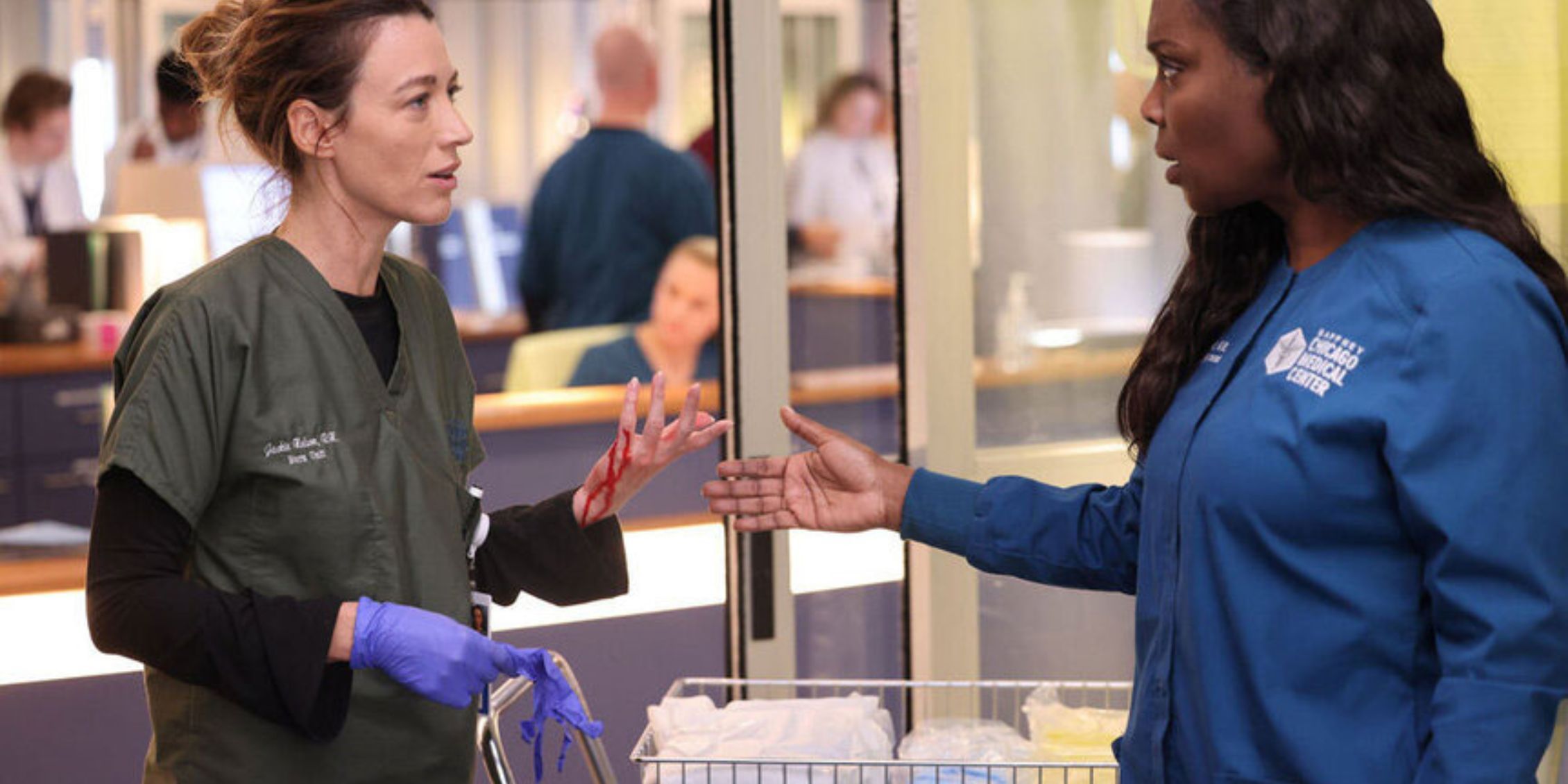 Chicago Med Jackie stands with a cart and a bloody hand while Maggie holds out her hand to make a point