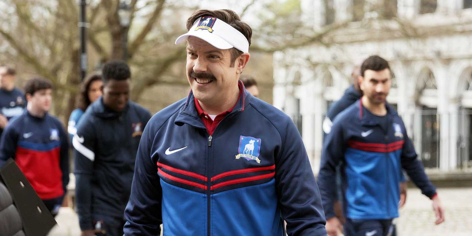 Jason Sudeikis as Ted Lasso smiling while the team walks behind him in Ted Lasso season 3, episode 1