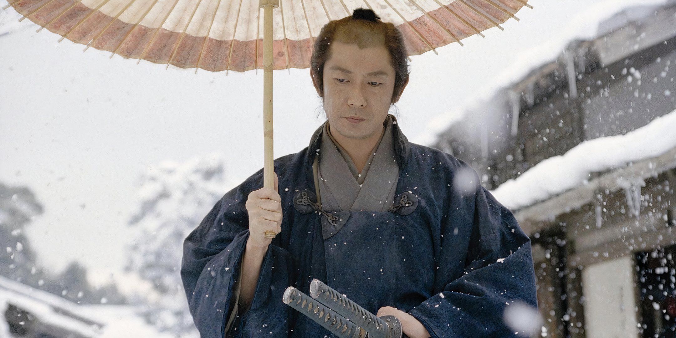 Masatoshi Nagase walks through the snow while holding a paper umbrella over his head.