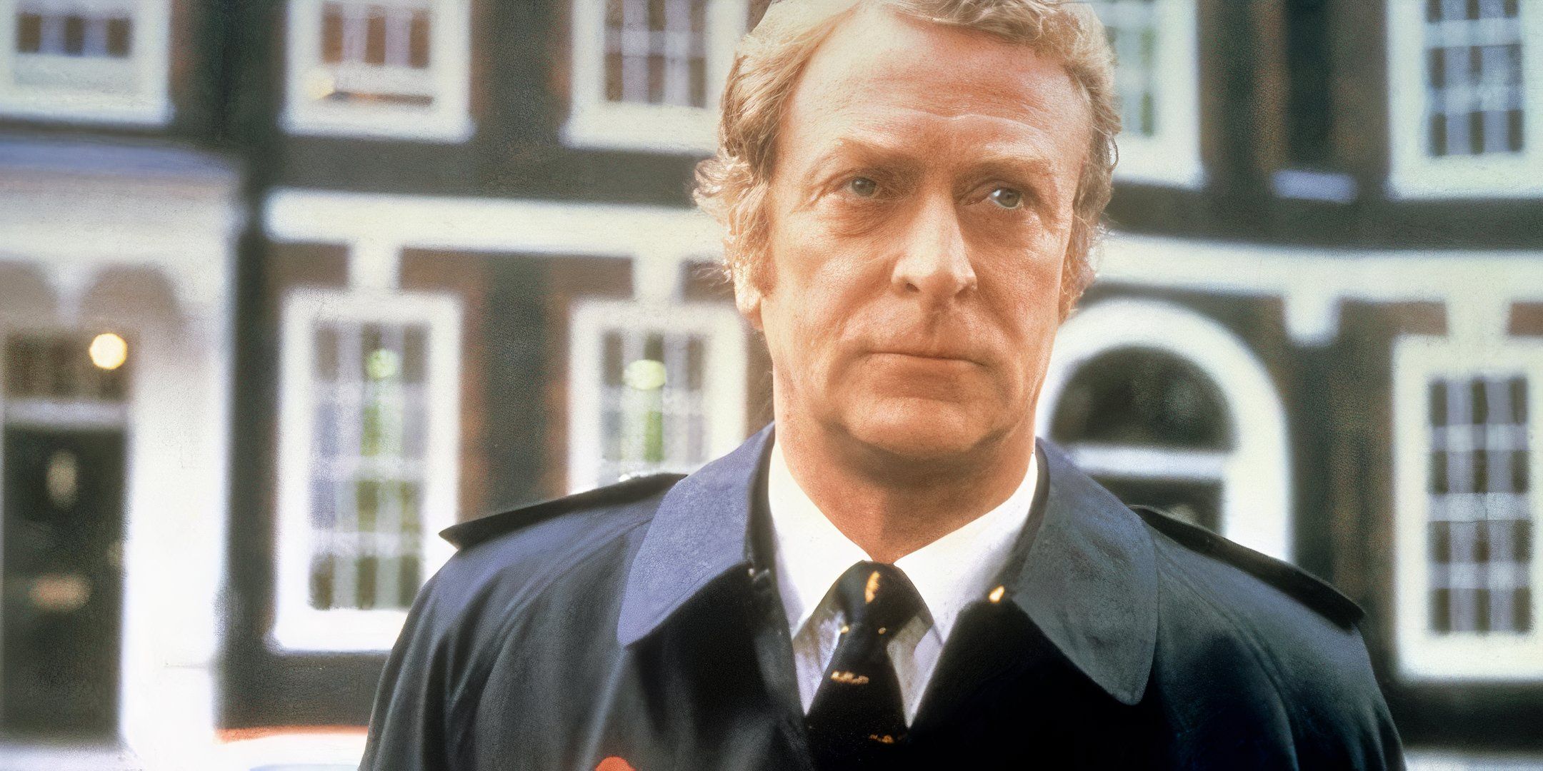 Michael Caine wears a uniform and stands outside, looking straight ahead. 