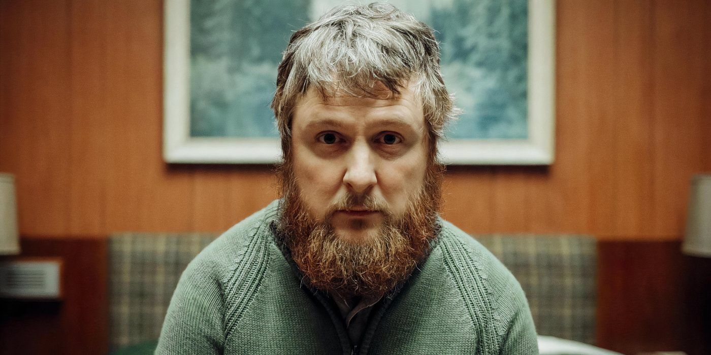 Tim Key sits on the bed and looks towards the camera