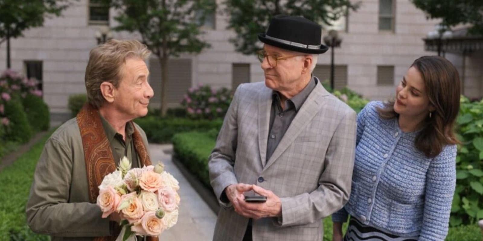 Oliver, holding flowers, looks at Charles and Mabel, who are turned towards him, in the Arconia courtyard in the Only Murders in the Building season 4 finale