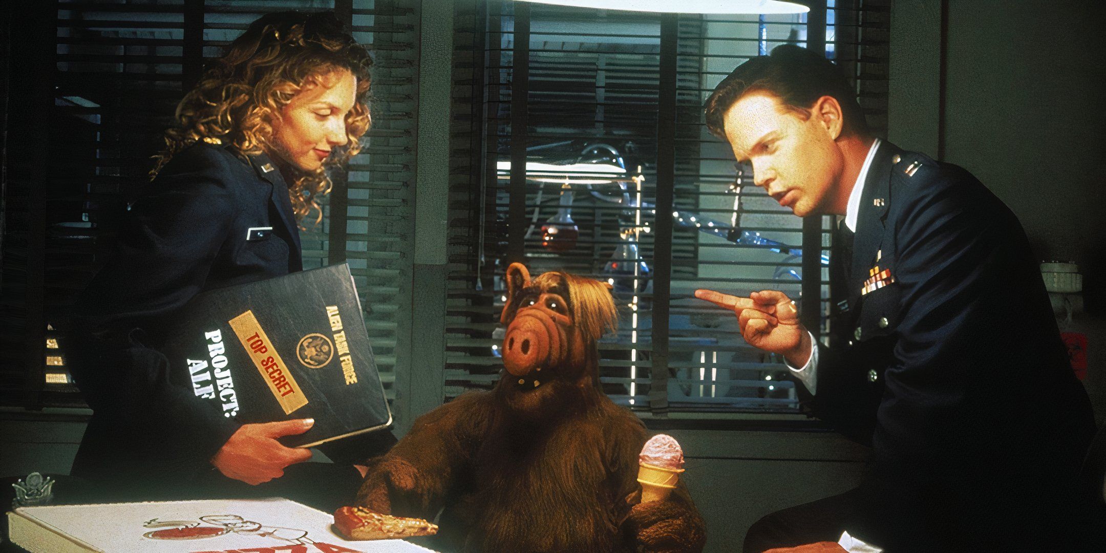 ALF sits at a desk while holding an ice cream cone and is being interrogated by government officials. 