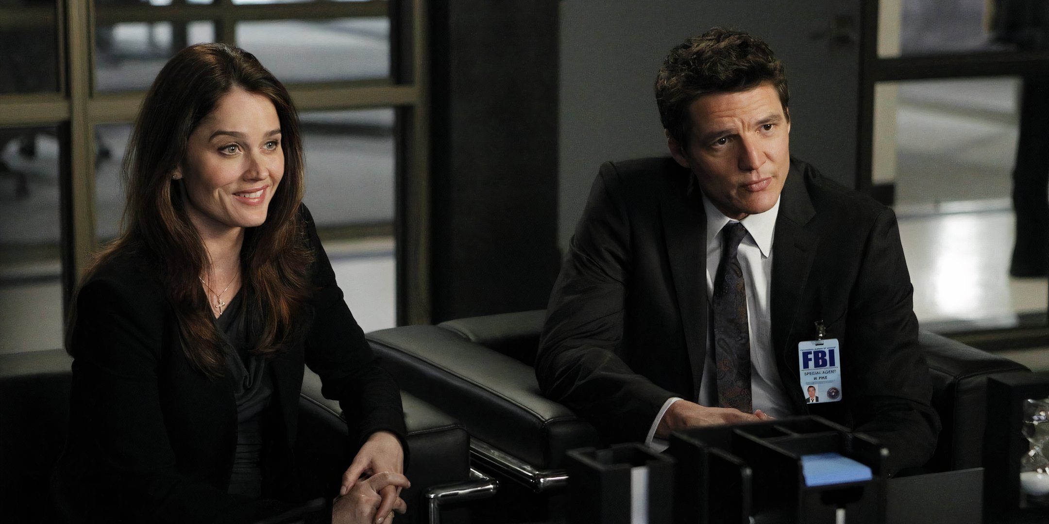 Robin Tunney and Pedro Pascal smile while sitting in front of someone's desk. 