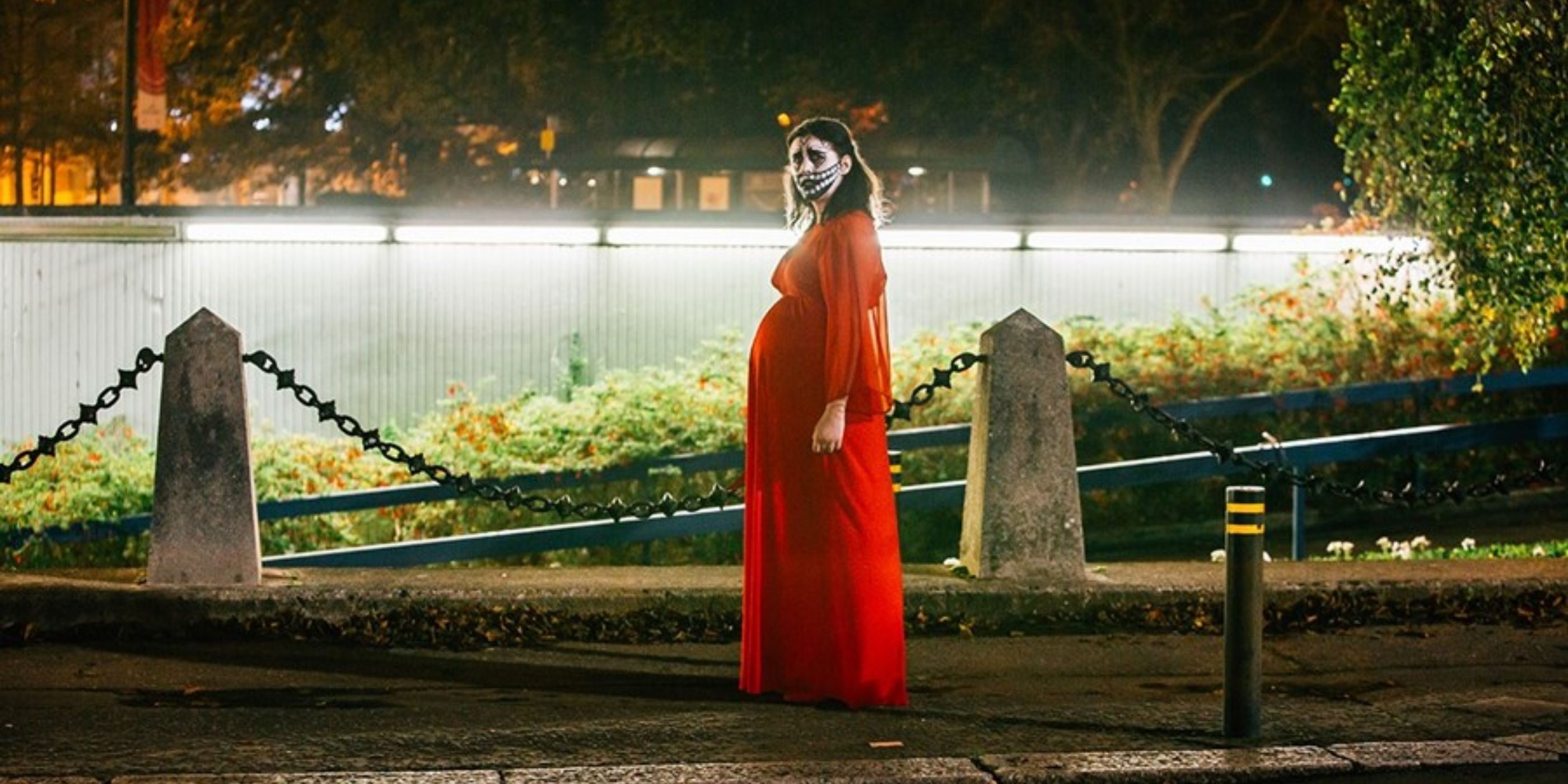 Ruth played by Alice Lowe in Prevenge in a red dress and creepy black and white makeup stands on the street staring at the camera