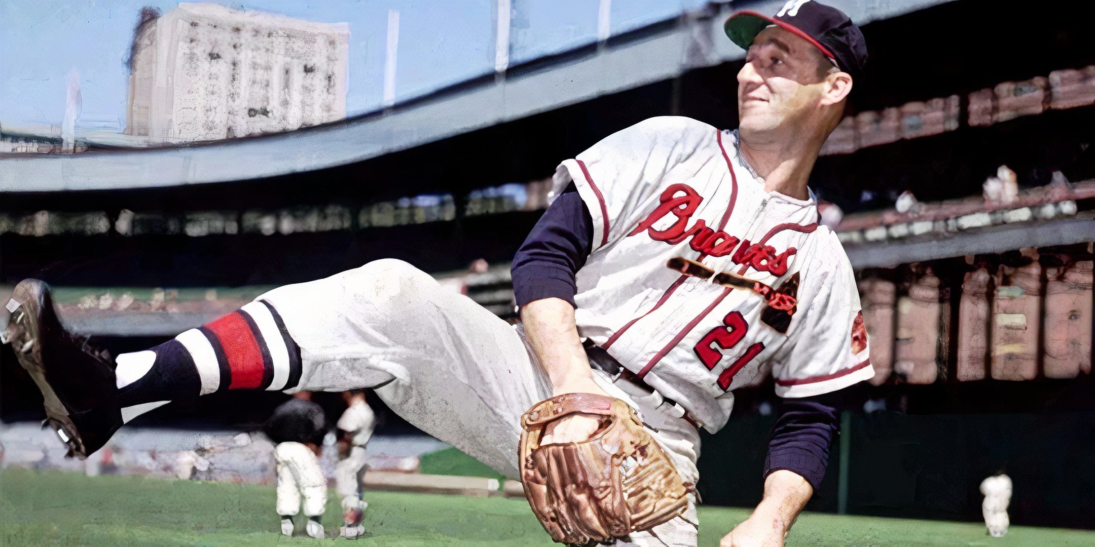 Uma fotografia colorida do lendário arremessador de beisebol Warren Spahn, vestindo seu uniforme do Boston Braves, no meio do campo em um estádio clássico. Spahn é capturado com a perna levantada e a luva posicionada, mostrando sua icônica forma de corda. 