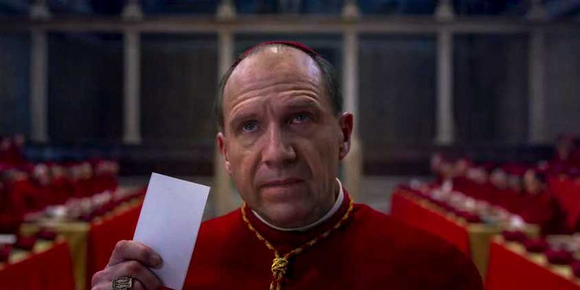 Cardinal Lawrence holding up a slip of paper while looking upwards in Conclave