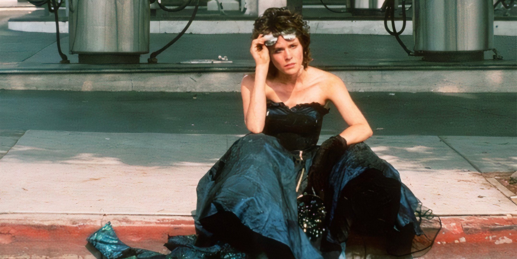 Ally Sheedy sits in a black dress on the curb. 