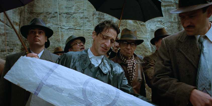 László showing his blueprint to a huddle of men with umbrellas in The Brutalist