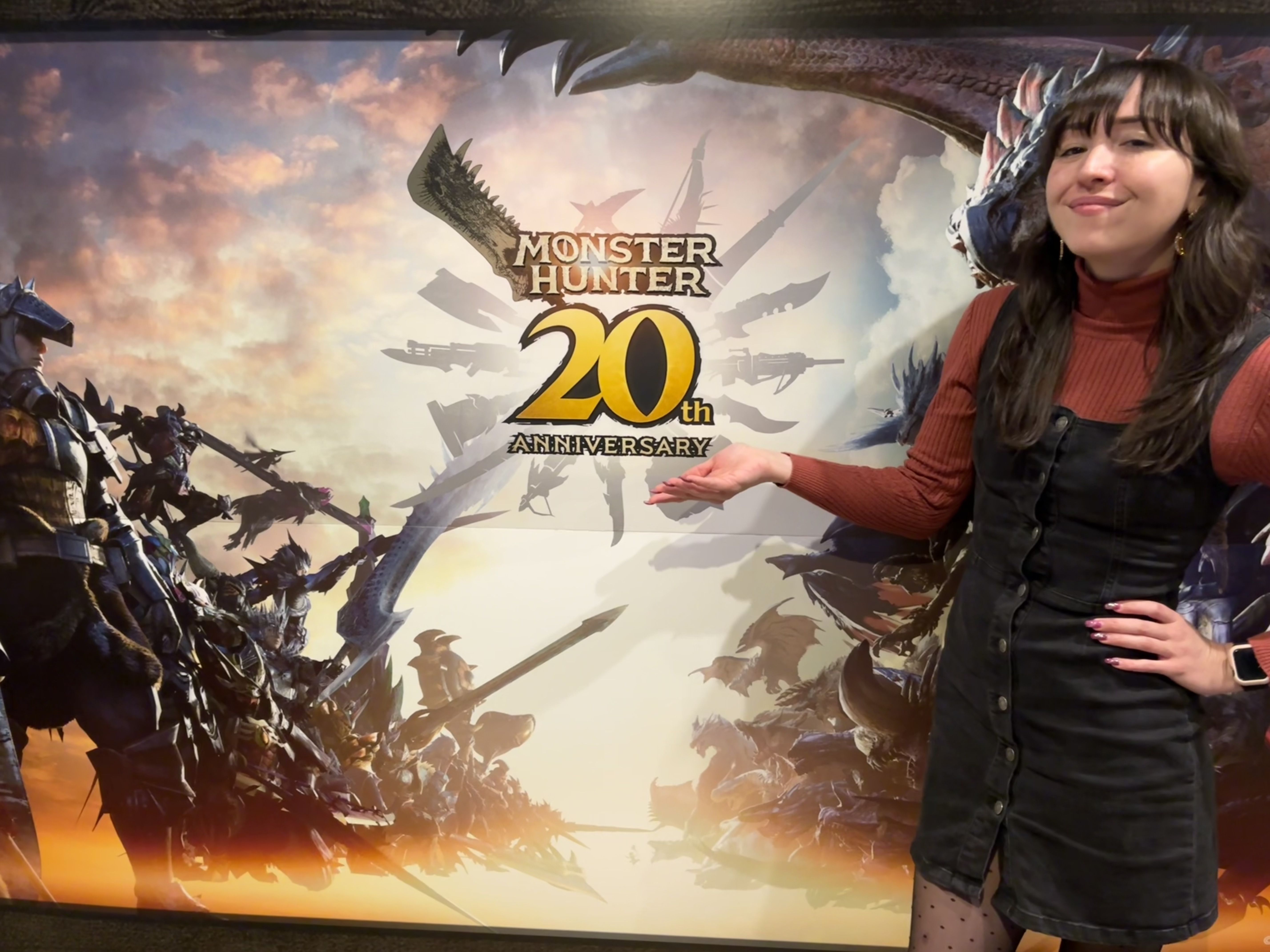 Woman standing in front of and gesturing to a sign that says Monster Hunter 20th Anniversary.