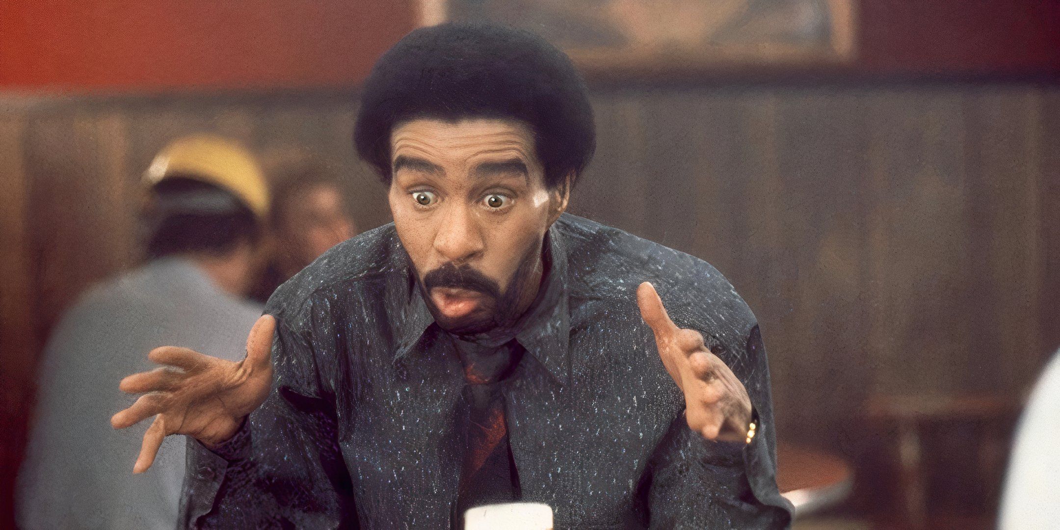 Richard Pryor sits at a table with a shocked expression on his face. 