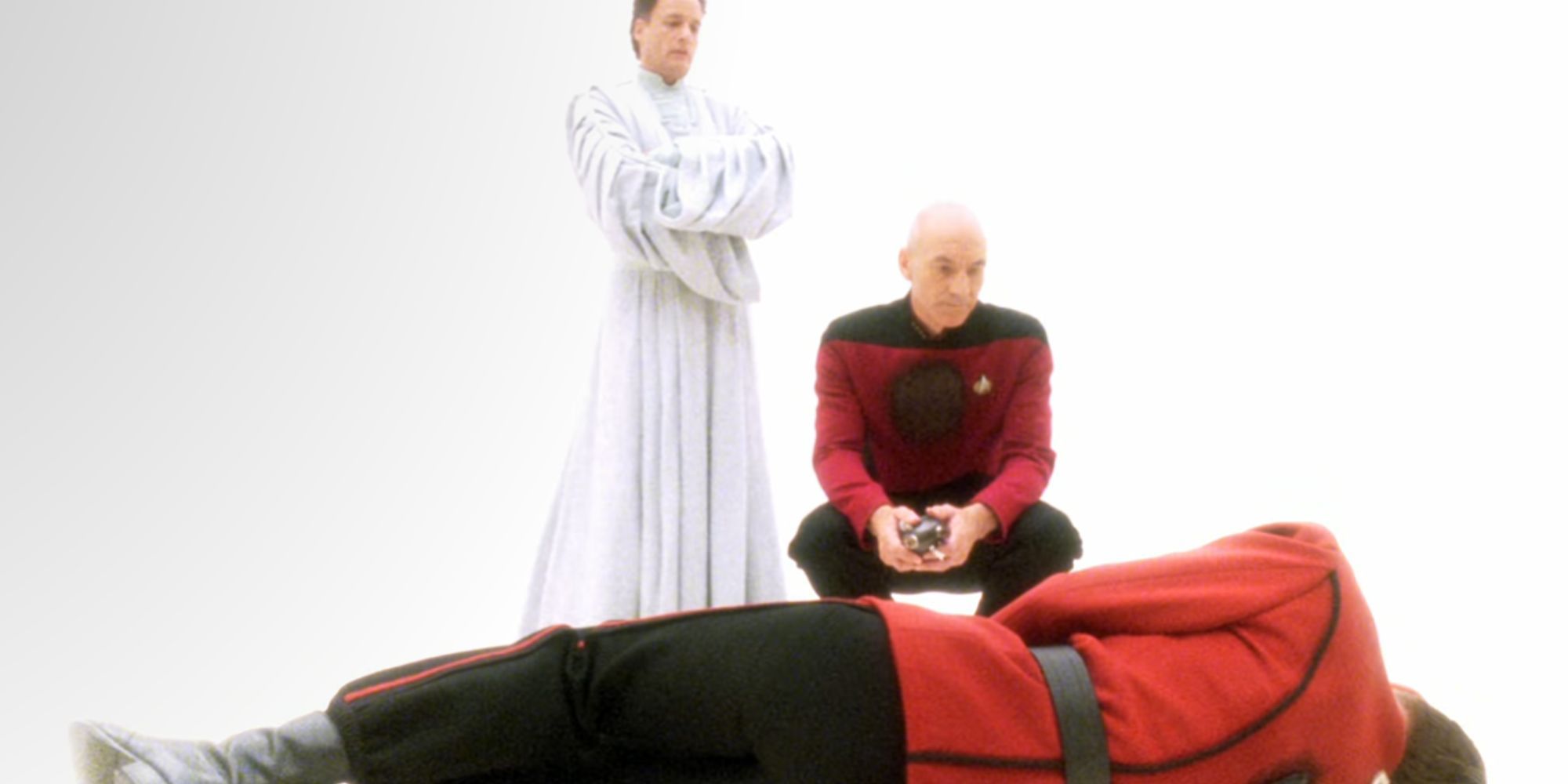 John de Lancie con una túnica blanca como Q en Star Trek: The Next Generation con Patrick Stewart agachado junto a él como el Capitán Picard mientras ambos miran al oficial caído de la Flota Estelar.