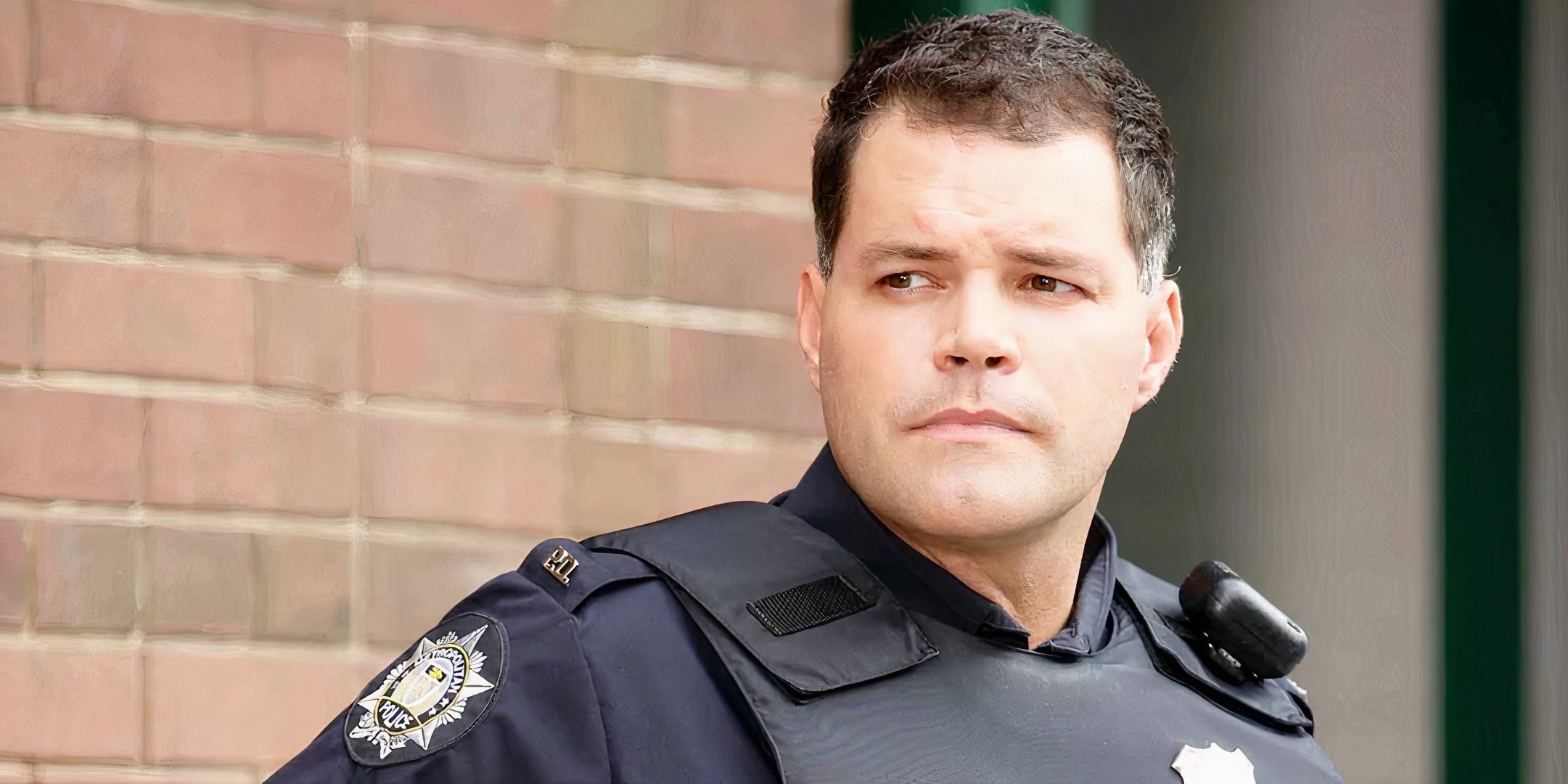 Aaron Douglas está vestindo uniforme de policial, parado em frente a uma parede de tijolos e olhando para a direita.