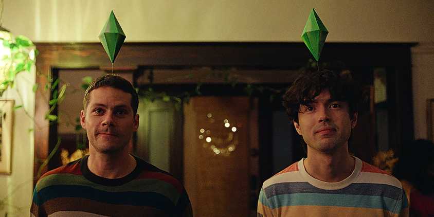 Dylan O'Brien and James Sweeney stand next to each other with Sims game icons over their heads