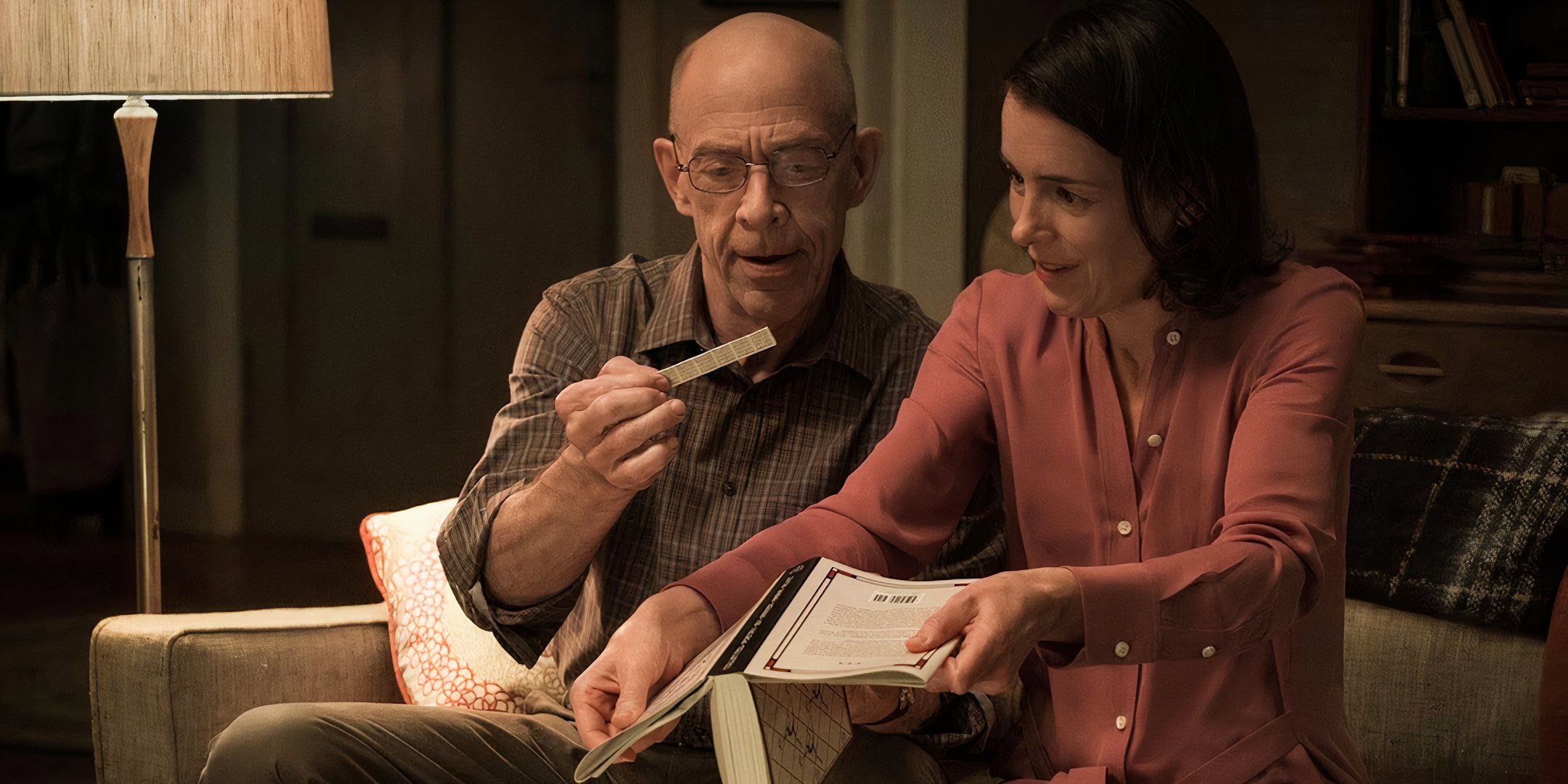 J. K. Simmons y Olivia Williams se sientan juntas en el sofá y examinan una hoja de papel.