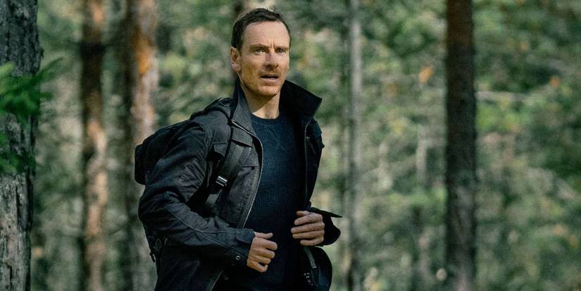 Michael Fassbender in a forest, wearing a thin black jacket in The Agency