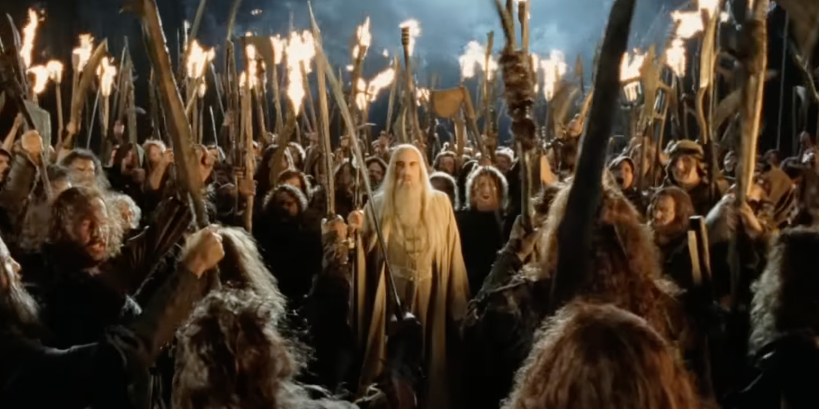 Saruman, a tall white man with a long beard and white robes, surrounded by a mob of angry white people in rags holding torches and crude weapons