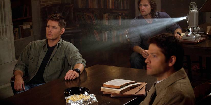 Castiel, Dean, and Sam watching a projector in the Supernatural episode "Clip Show." 