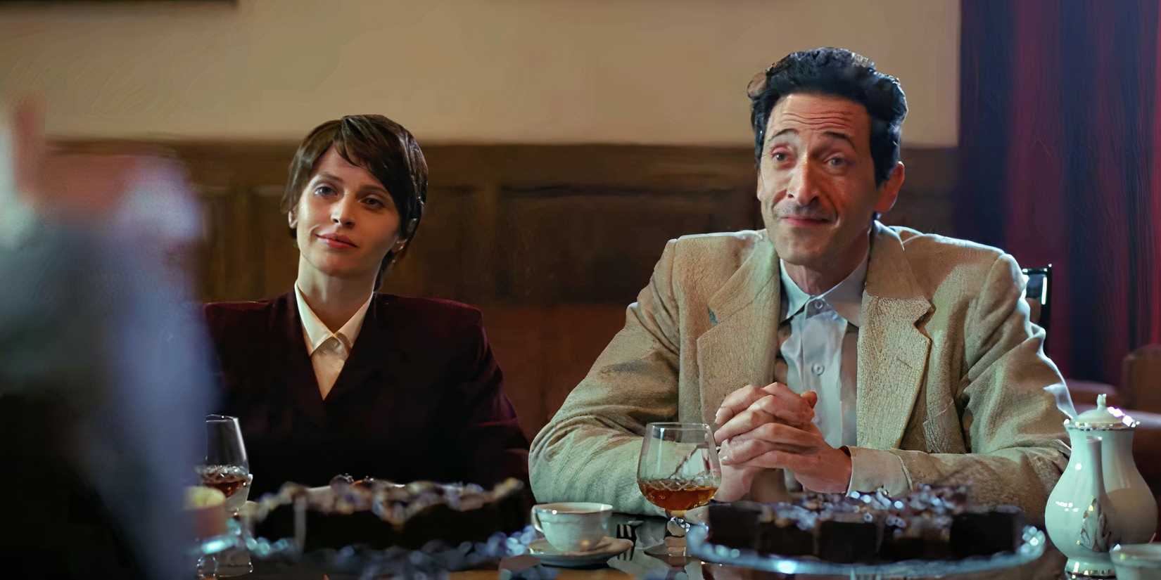 Adrien Brody and Felicity Jones sitting together at a dinner table in The Brutalist