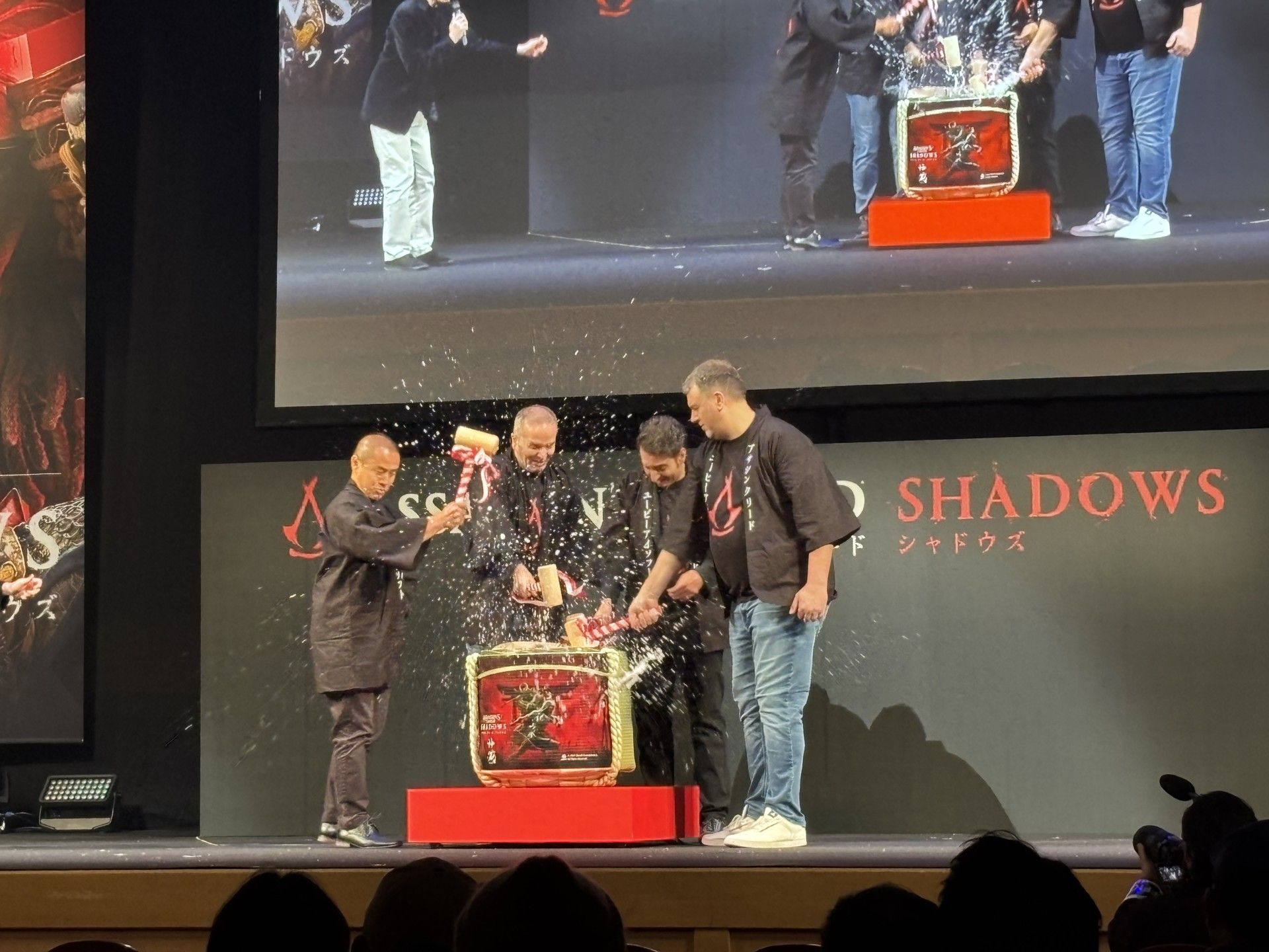 A group of four men on a stage smashing a sake cask with the Assassin's Creed Shadows art on it.