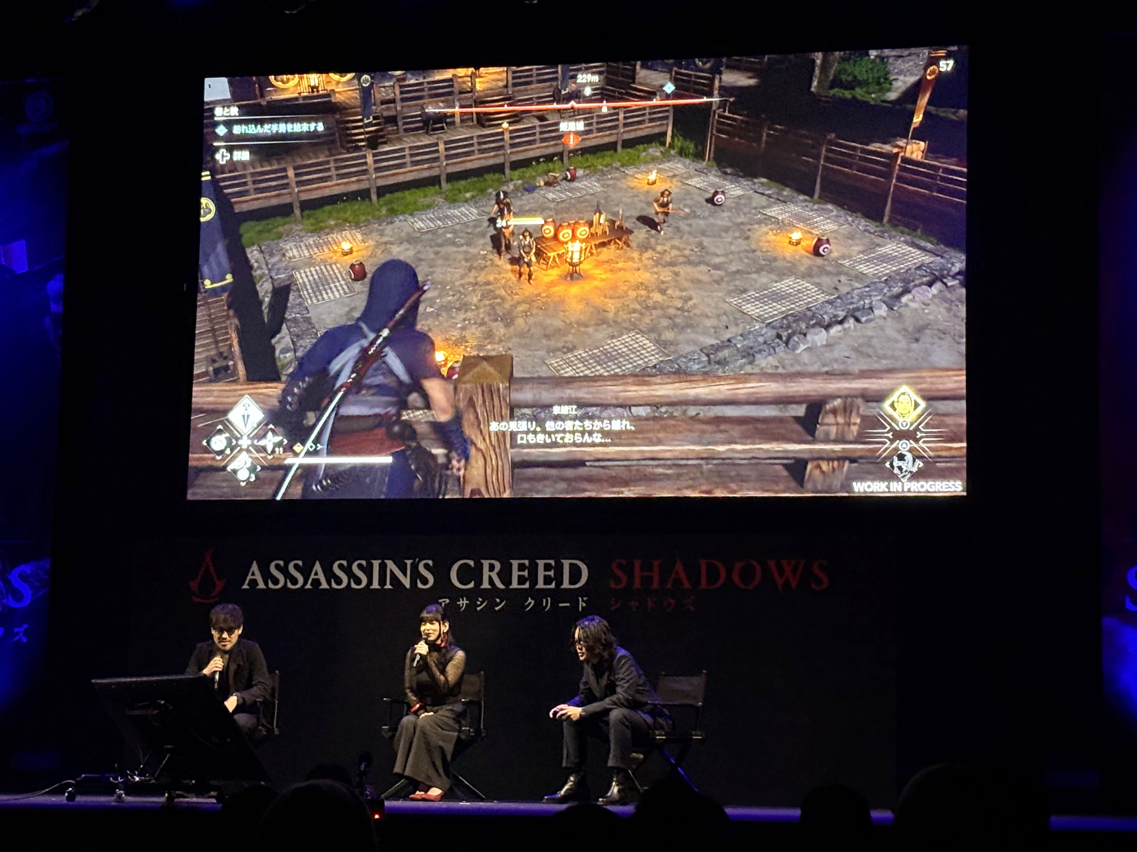 Two men and a woman sitting on stage as the one on the right plays Assassin's Creed Shadows.