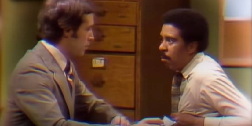 Chevy Chase sitting at a desk across from Richard Pryor on SNL