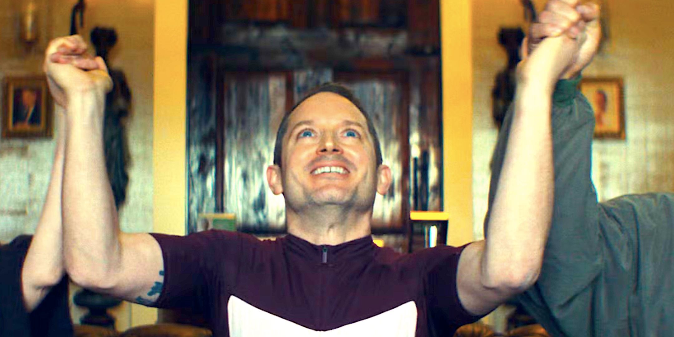 Elijah Wood&rsquo;s Ted sits on a couch holding two people&rsquo;s hands while smiling in The Monkey 2025