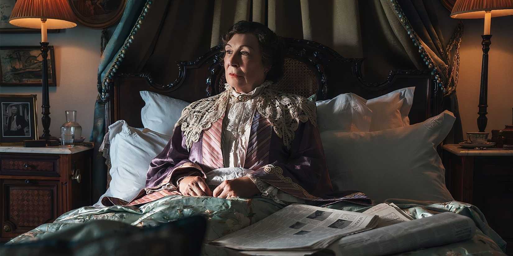 Lady Tressilian (Huston) sits in her Victorian bed looking solmenly to the sunlight while draped in lavish pajamas. A newspaper rests on top of the bed