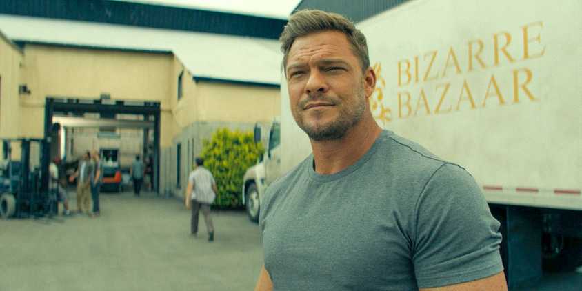 Alan Ritchson as Reacher standing outside next to a big 18-wheeler