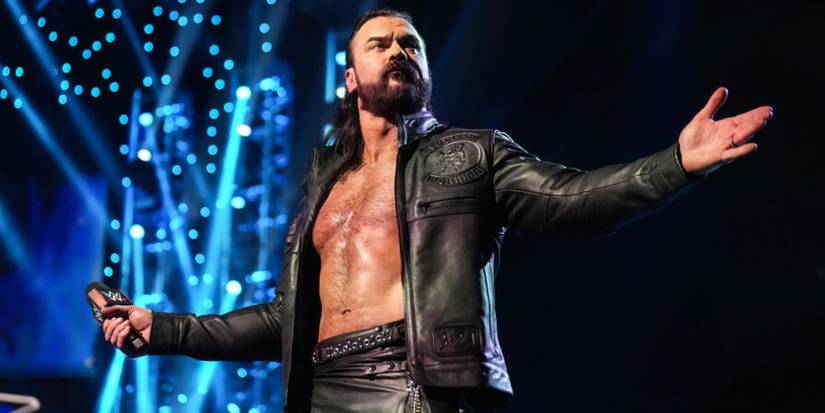 Drew McIntyre, microphone in hand, looks out the audience with his arm spread out. 