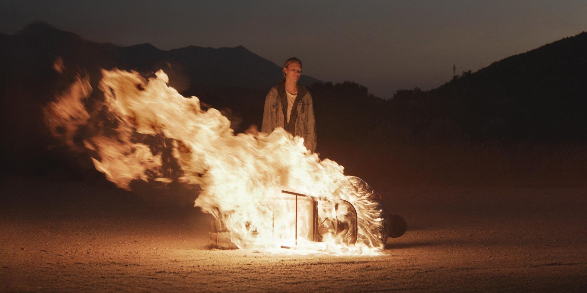 A woman stands in front a man as he burns alive in a chair in Redux Redux