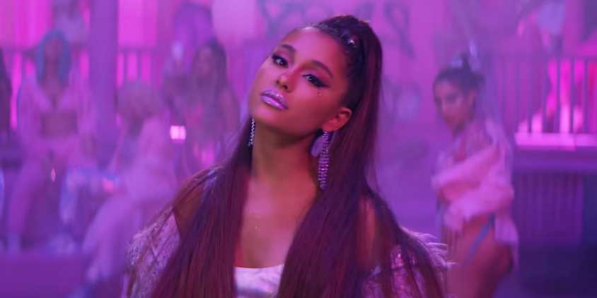ariana grande has on a white dress, her hair is in a high ponytail and comes down over her shoulders. She has silver dangling earrings, and silvery lipstick. The background is blurred, but it has a purple tint and some people are visible.