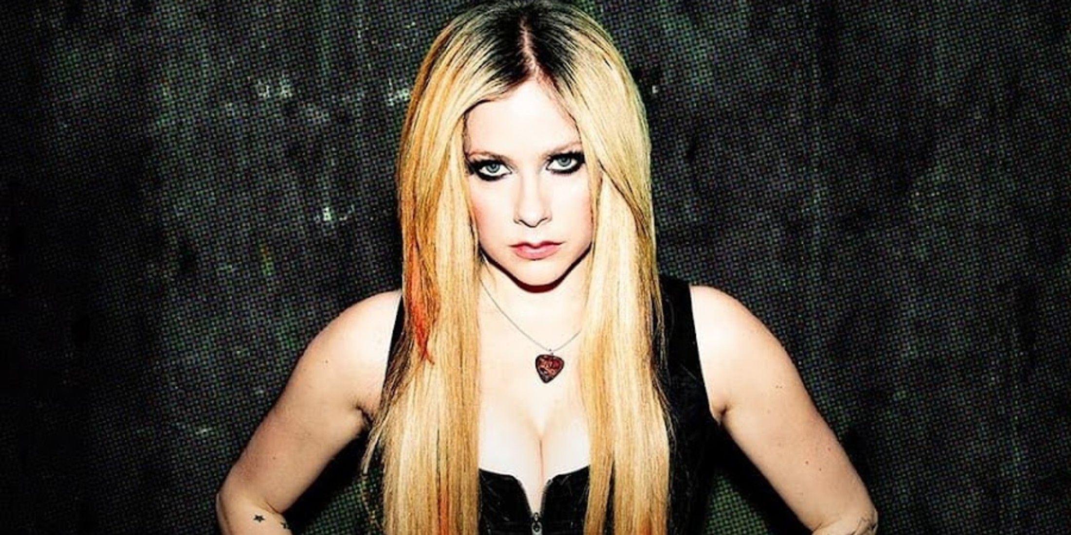 avril lavigne stands in front of a black and white background. She has blonde hair, wears a black top, and a necklace with a guitar pick on it.