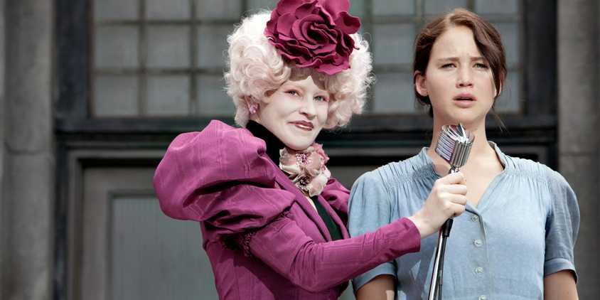 Effie Trinket (Elizabeth Banks) smiling while putting the microphone in front of Katniss Everdeen (Jennifer Lawrence) in The Hunger Games