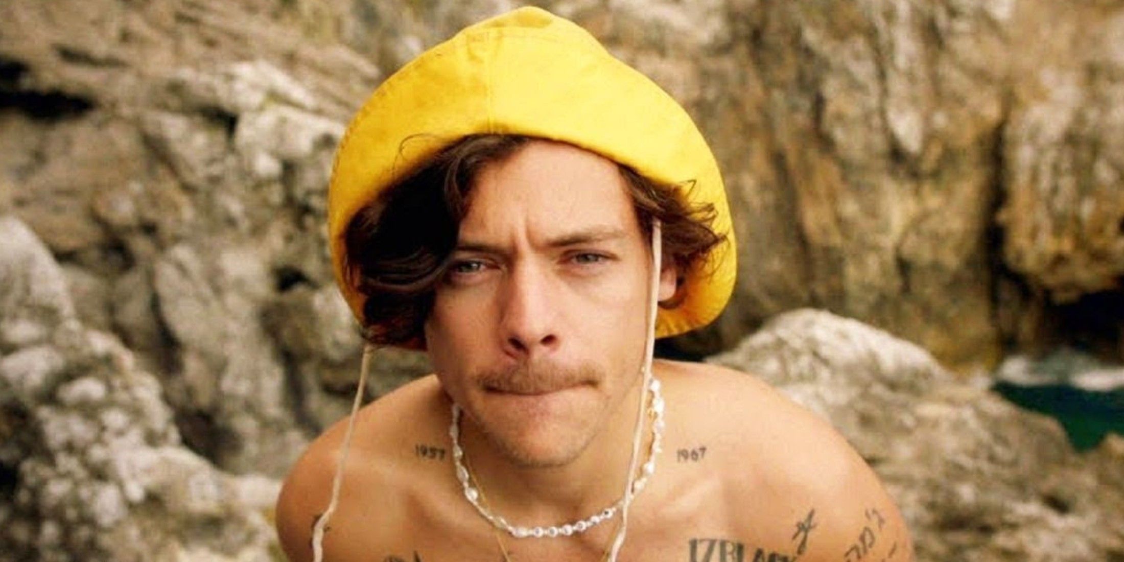 harry styles with rocks in the background. He is shirtless with a white necklace and a yellow cap on.