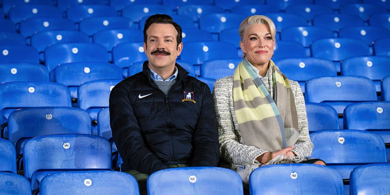 Jason Sudeikis as Ted and Hannah Waddingham as Rebecca sitting in the blue seats of Richmond Stadium in Ted Lasso.