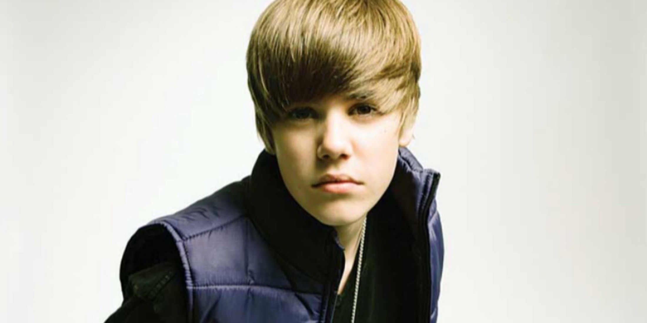 justin bieber looking into the camera, wearing a vest with his signature swooped hair style, in front of a white background.