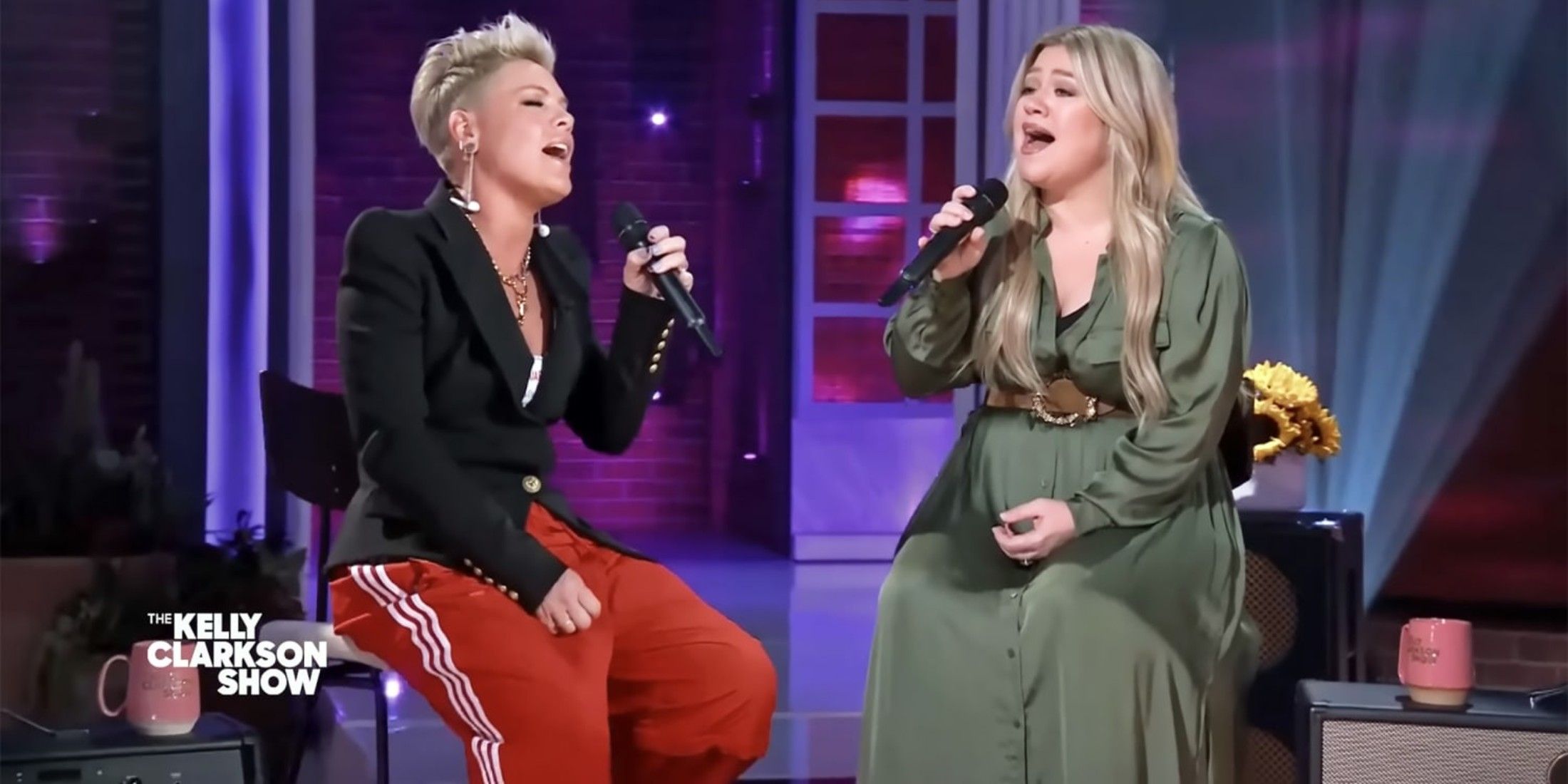 Pink sits in a chair holding a microphone, while wearing a black coat and red pants with three white strips on the side. Kelly Clarkson sits next to her holding a microphone and wearing a green dress.