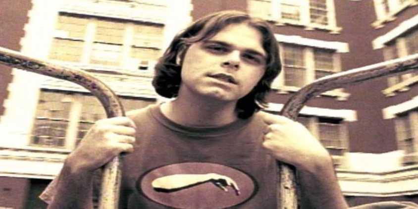 a grainy photo with a person gripping two bars. A building is in the background. The person has hair to his shoulder and a shirt with a logo that has a forearm on it. 