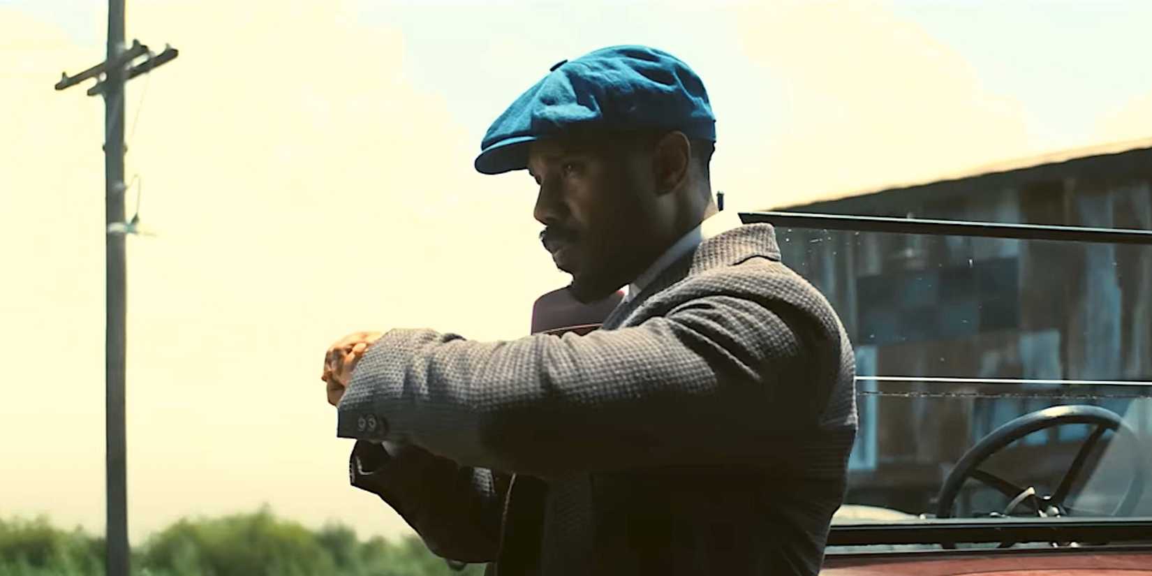 Michael B Jordan's character looks at his watch while standing by a car in Sinners