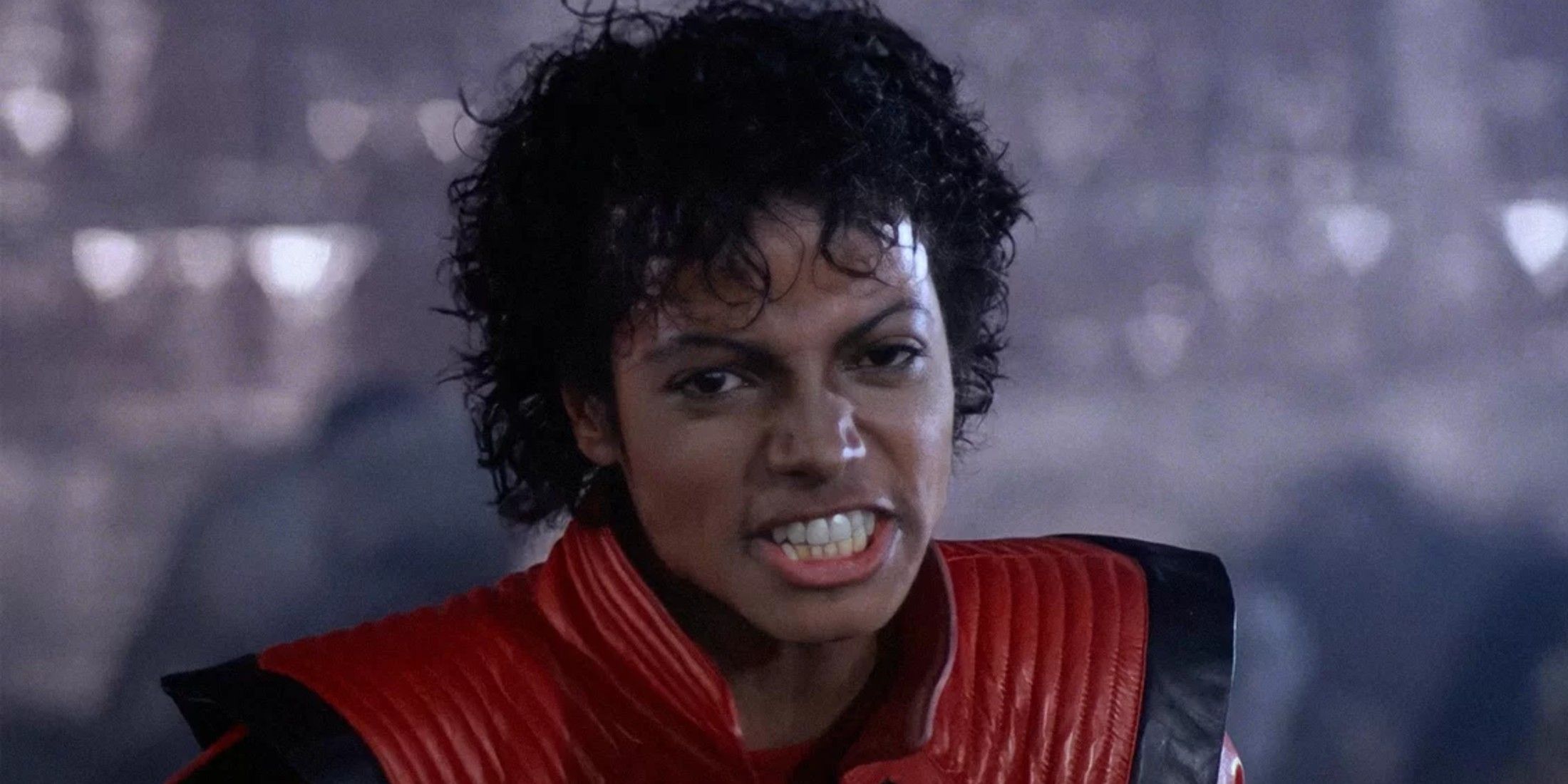 Michael Jackson stands in front of a blurred background wearing a red and black coat, he is showing his teeth and has medium dark curly hair.