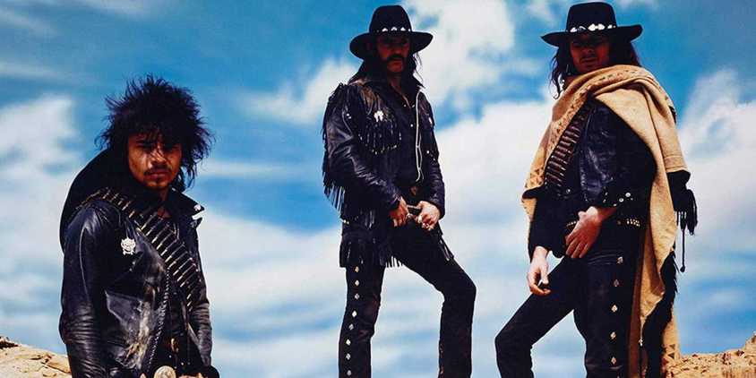 The background is a blue sky mostly covered by white cloud. There are three people the one wears a black leather jacket, has a goatee mustache and has long black hair. The one in the middle has on a black leather jacket, and black leather pants. He has on a black cowboy hat and his black hair is visible. The one of the far right has on black leather pants and a black leather jacket. He has on a tan pancho that covers come of the jacket. His long black hair sticks out from his black cowboy hat.