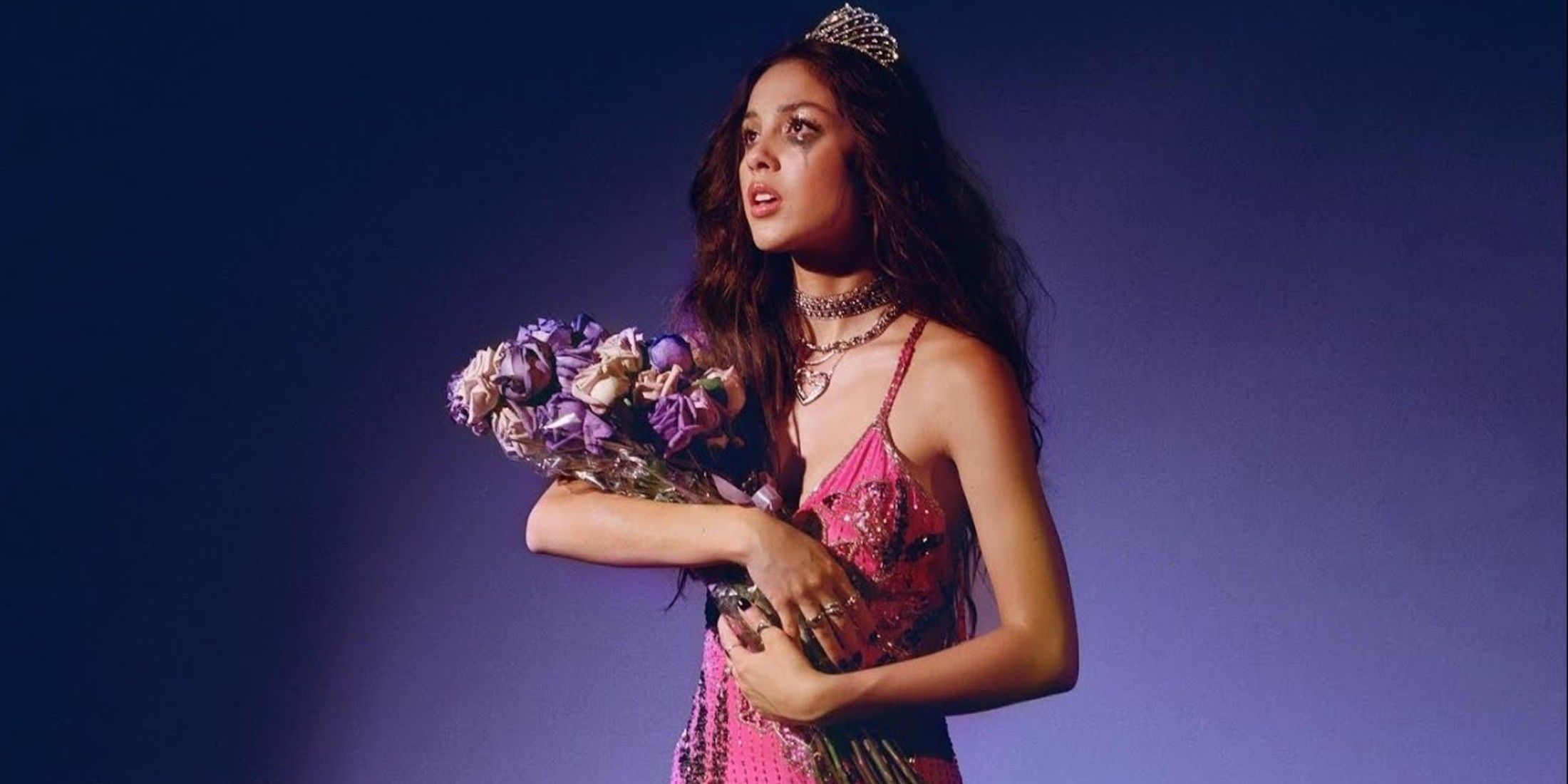Olivia Rodrigo stands in front of purple background. She holds white and purple roses. She is wearing a pink dress with black flowers on it. She has on a sliver crown on top of her long dark hair. Her makeup is running to show crying.