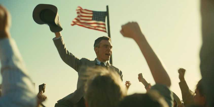 Oppenheimer triumphantly raising his hat to a cheering crowd while the American flag waves behind him